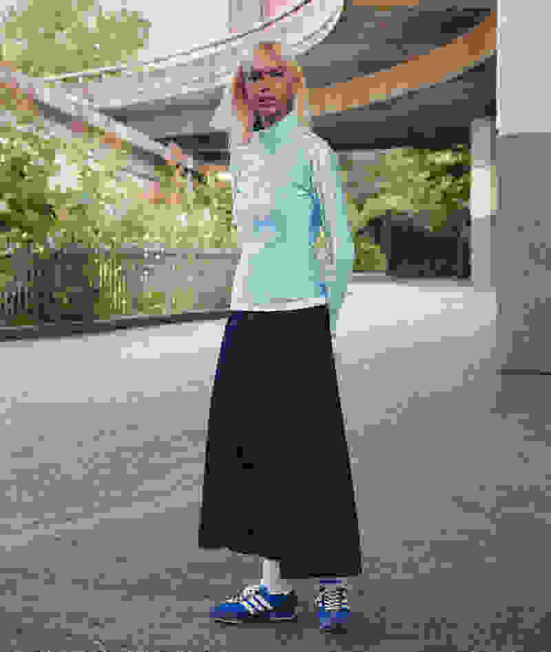 A young woman with blonde hair stands in front of a curved concrete structure, wearing a light blue top and a long navy blue skirt, paired with blue sneakers.