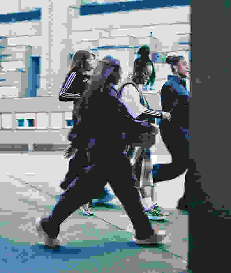 A group of people, some wearing athletic clothing, are walking quickly through an urban setting with buildings and structures in the background.