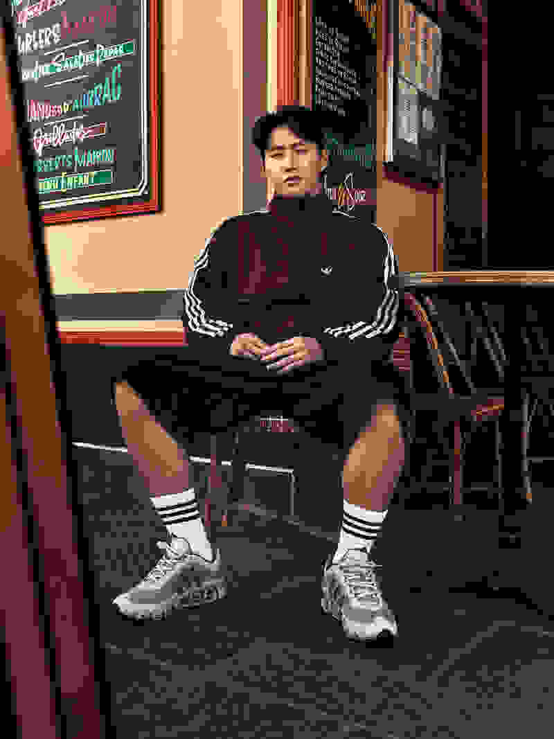 A young man wearing a burgundy jacket and black shorts sits on a chair in front of a storefront with various signs and advertisements displayed in the background.