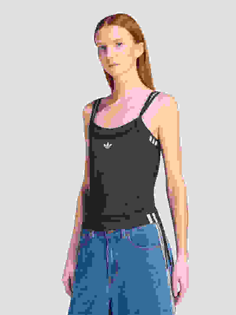 A young woman with long red hair is wearing a black tank top and blue jeans, standing against a plain white background.