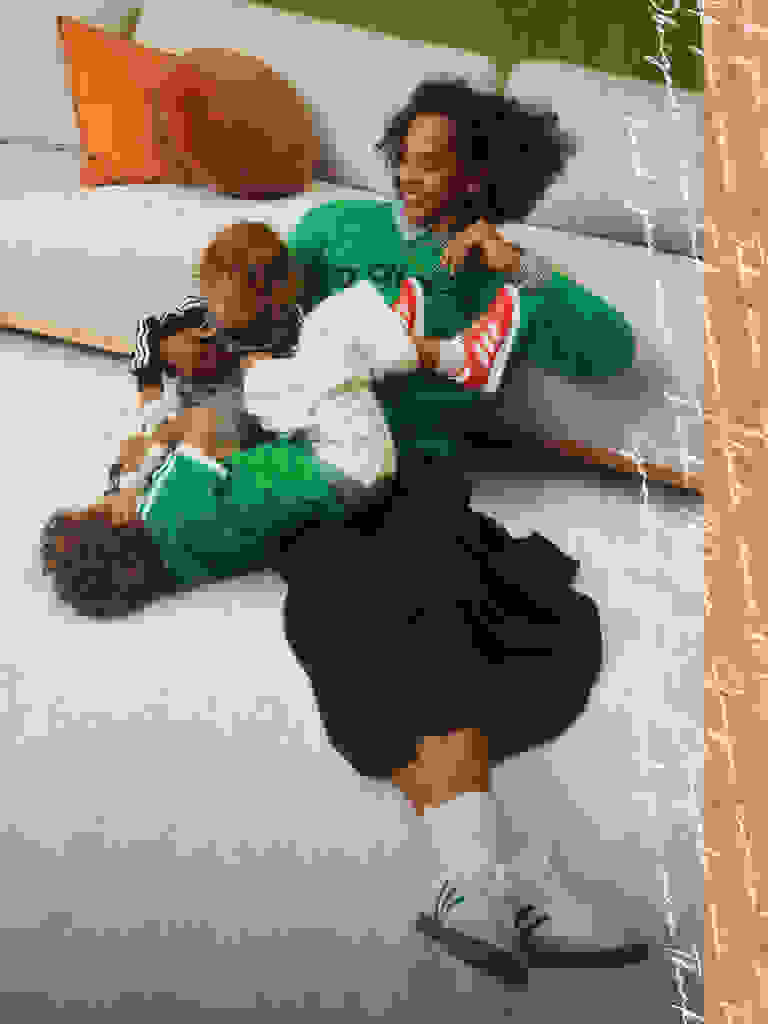 A woman with curly hair is lying on the floor, cuddling and playing with a young child in a cozy, home-like setting with colorful pillows and a brick wall in the background.