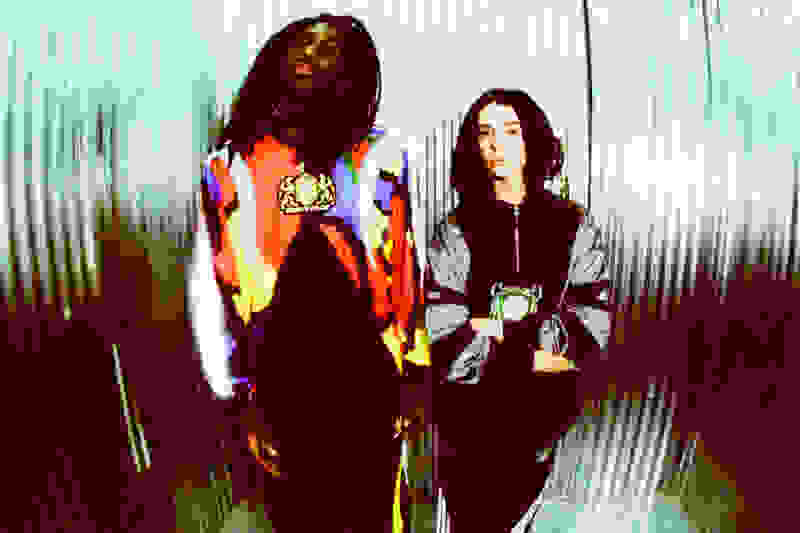 Two individuals, a person with long dark hair and another with shorter dark hair, stand in front of a blurred, reflective background.