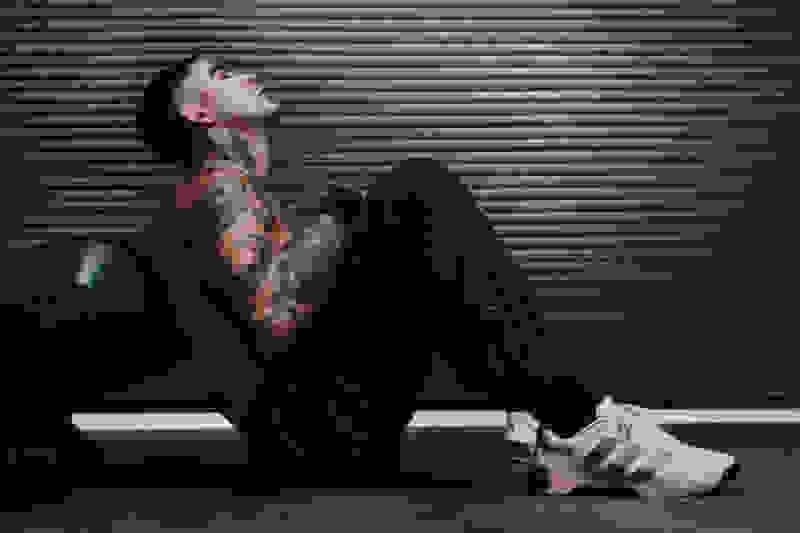 A person with tattoos sits on the ground in front of a metal shutter, wearing dark clothing and white sneakers.