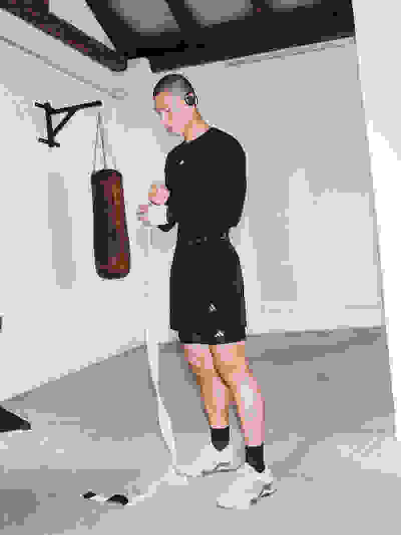 A person in black athletic clothing stands in a room with wooden beams and a punching bag, appearing to be preparing for a workout.