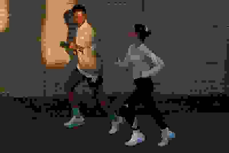 Two individuals, a man and a woman, are walking together at night against a backdrop of a brick wall.