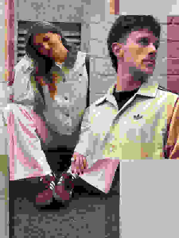 Two individuals, a young woman and a young man, are sitting on the ground in front of a brick wall. The woman is wearing a light-colored denim jacket and the man is wearing a beige jacket. They both appear to be casually dressed.