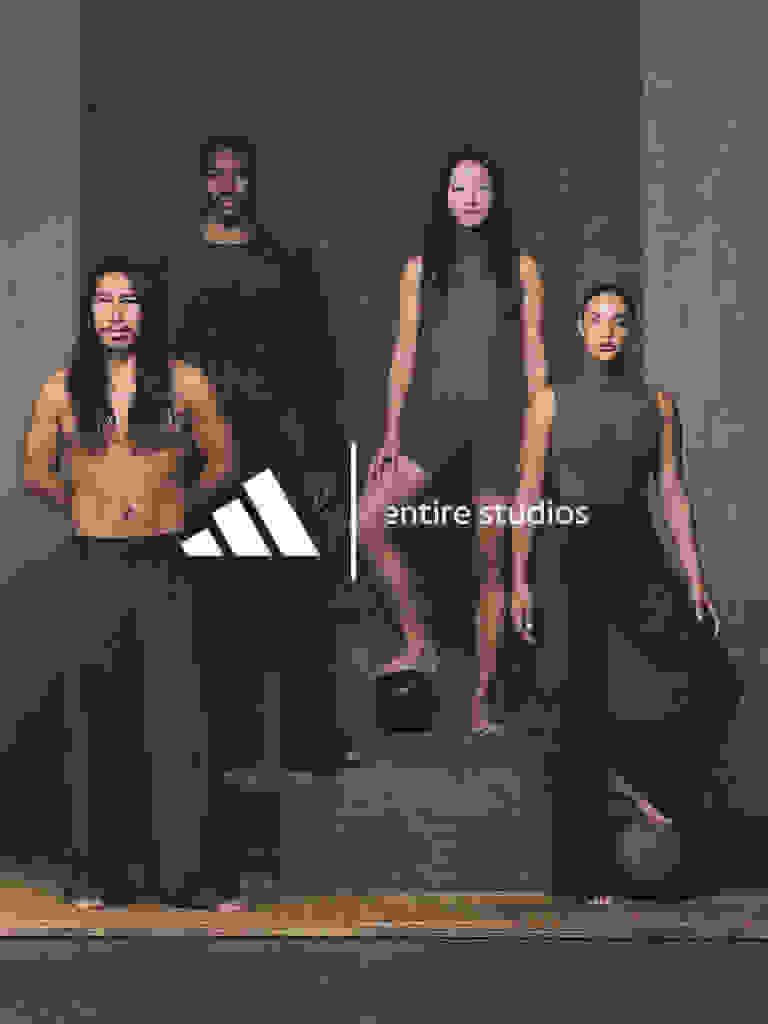 The image features a group of individuals, both male and female, posing together in a minimalist studio setting. The individuals are wearing dark-colored clothing, and the background is a plain, neutral-toned wall, creating a stark and dramatic visual contrast.