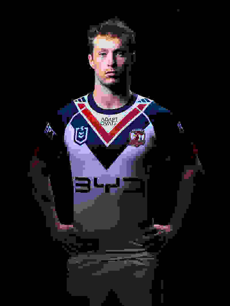 The image shows a young man wearing a sports jersey with the team's logo and sponsor prominently displayed, standing against a dark background.