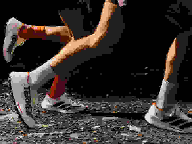 The image shows the lower body and legs of a person wearing white and orange athletic shoes, standing on a surface covered in small rocks or gravel.