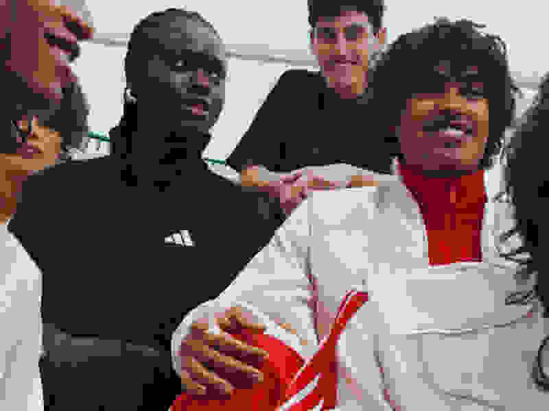 A group of young people, some wearing athletic clothing, are standing together and smiling in an outdoor setting with a clear sky in the background.