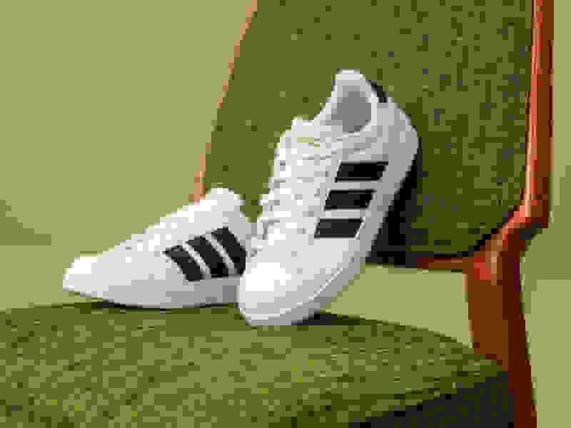 A pair of white Adidas sneakers with black stripes sits on a green upholstered chair against a green background.