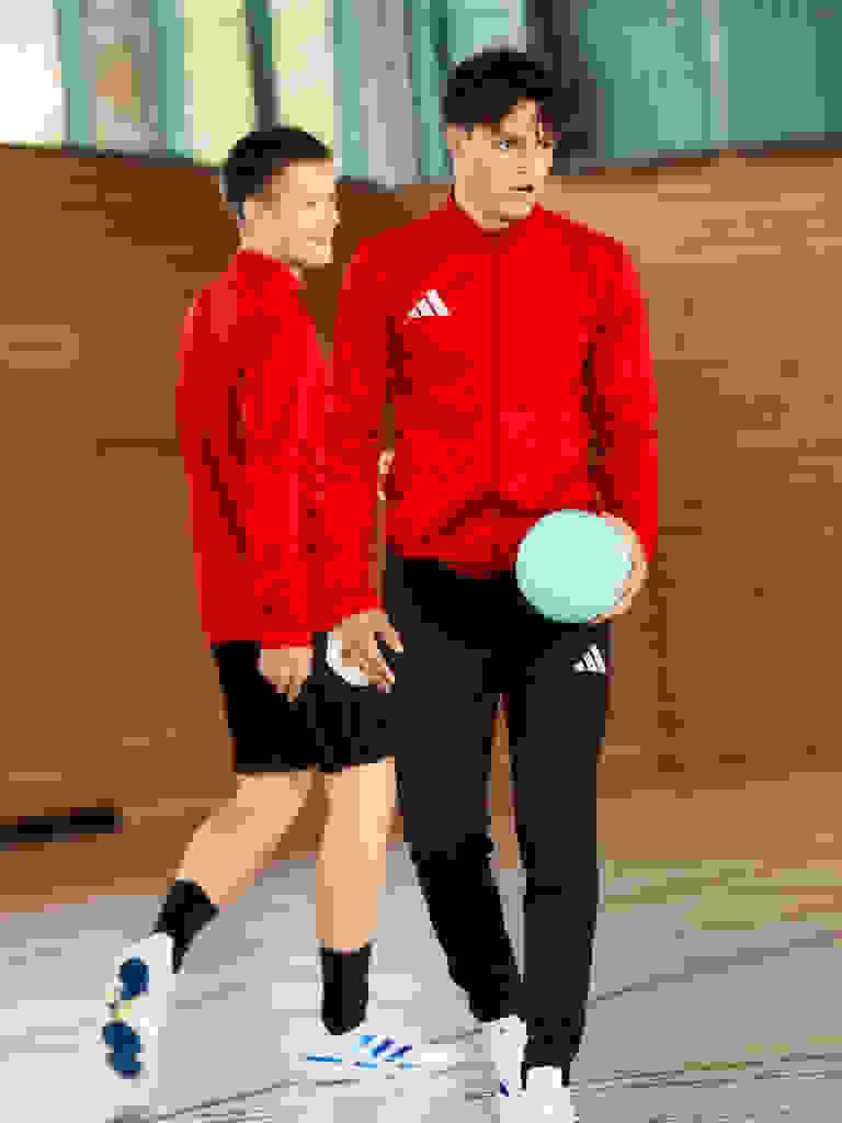 Two young boys wearing red Adidas jerseys and black shorts are standing on a wooden floor in what appears to be an indoor sports facility.