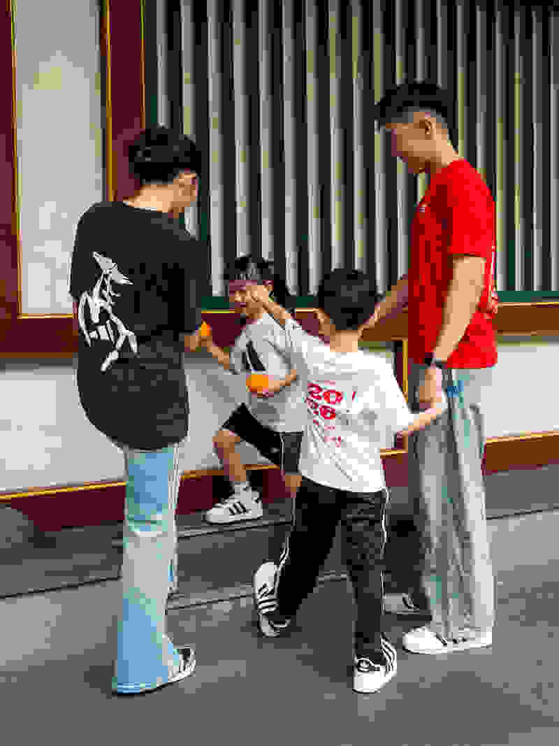 Four young individuals, dressed in casual attire, are engaged in a playful interaction on what appears to be a paved surface in front of a building with vertical blinds.