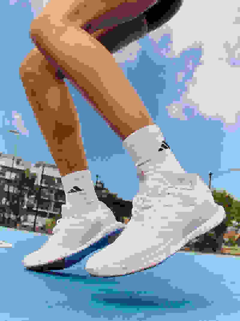 The image shows a person's legs and feet wearing white athletic shoes, standing on a tennis court with a blue sky and clouds in the background.