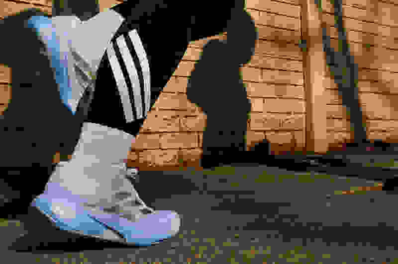 The image shows a pair of white and blue athletic shoes with three stripes on the side, standing on a concrete surface in front of a brick wall.