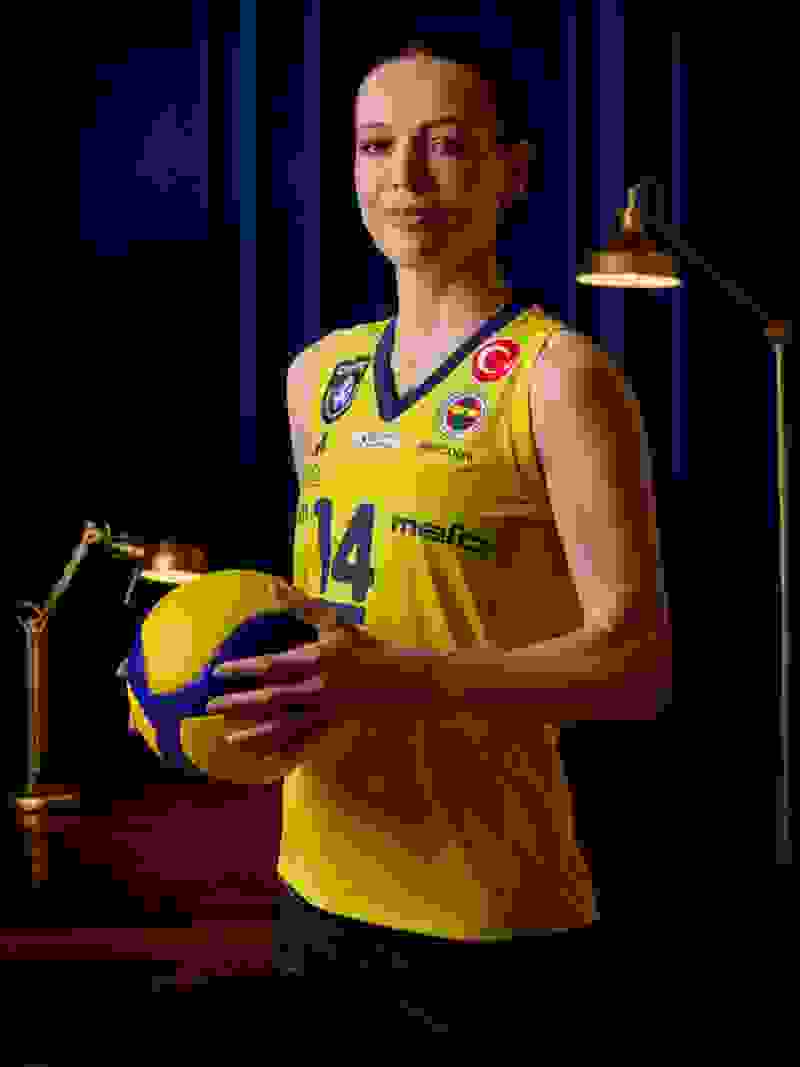 A young woman in a yellow volleyball jersey stands in a dimly lit room, holding a volleyball and smiling confidently.