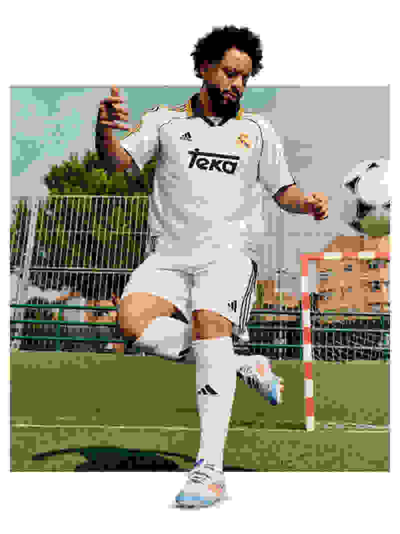 A soccer player in a white jersey and shorts is standing on a soccer field, with a soccer ball at his feet and a goal post visible in the background.