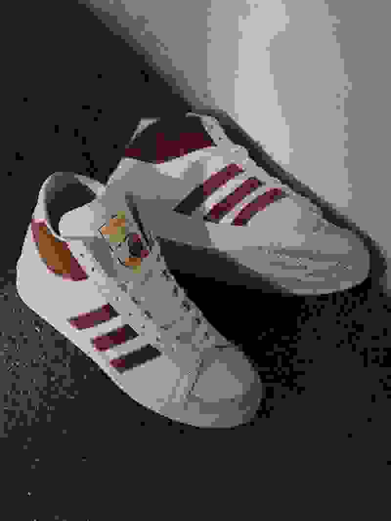A pair of white and maroon sneakers with the Adidas logo are shown on a dark surface.