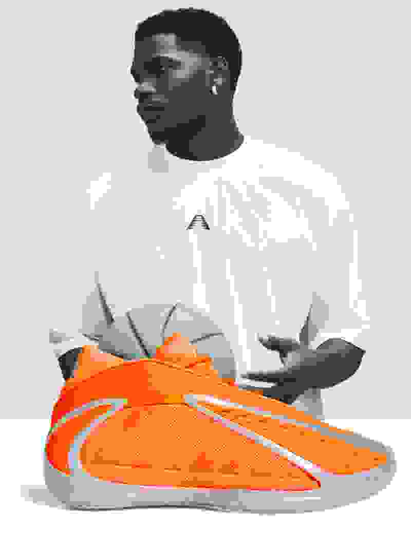 The image features a person wearing a white t-shirt and holding an orange and white athletic shoe in the foreground, with a plain white background.