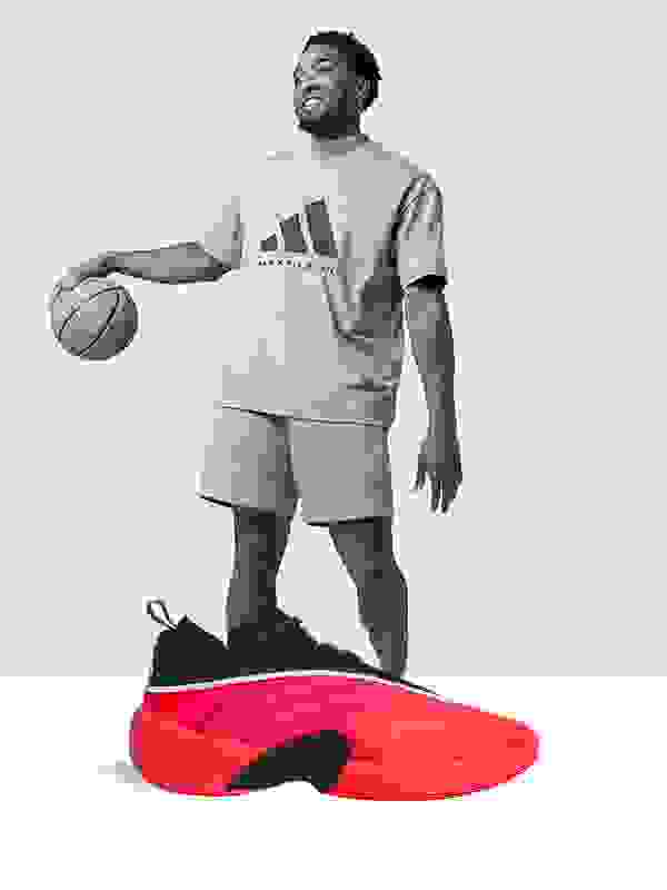 A smiling man wearing an Adidas t-shirt and shorts stands next to a large red basketball shoe in the foreground.