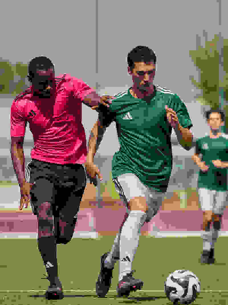 Two soccer players, one in a pink jersey and the other in a green jersey, are engaged in a competitive play on a soccer field with spectators in the background.