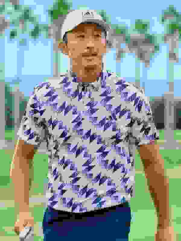 A man wearing a white cap and a patterned blue and white shirt stands on a grassy field with palm trees in the background.