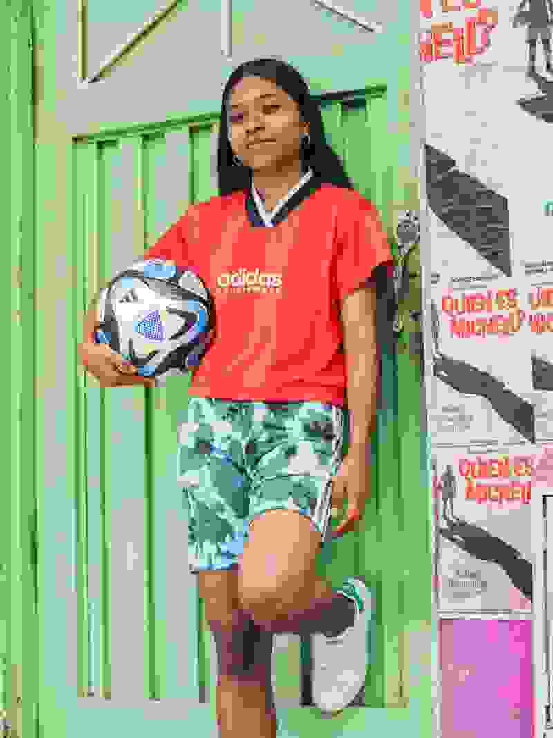 A young woman in a red Adidas jersey is holding a soccer ball, standing in front of a green wooden door with posters on it.