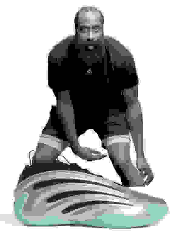 A large, bearded man in a black shirt is crouched down and examining a pair of futuristic-looking sneakers with a distinctive wavy sole design.