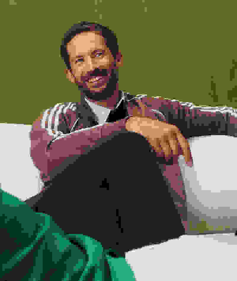A smiling man with a beard sits comfortably on a couch, wearing a striped shirt and dark pants against a green background.