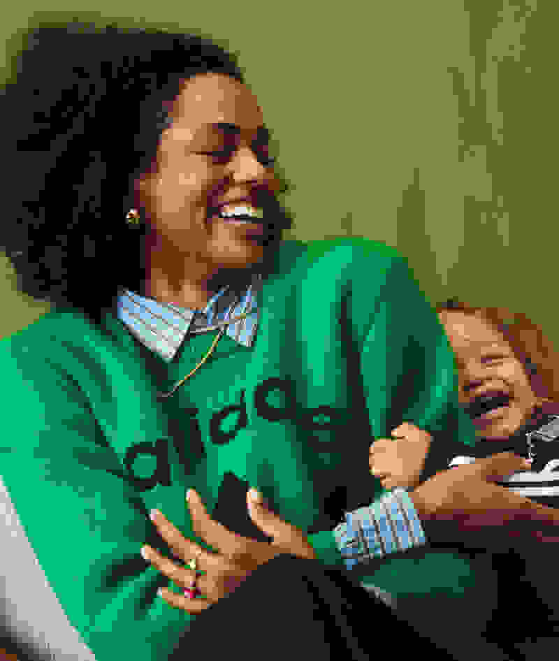 A smiling woman in a bright green sweater embracing a young child, both appearing joyful against a plain green background.