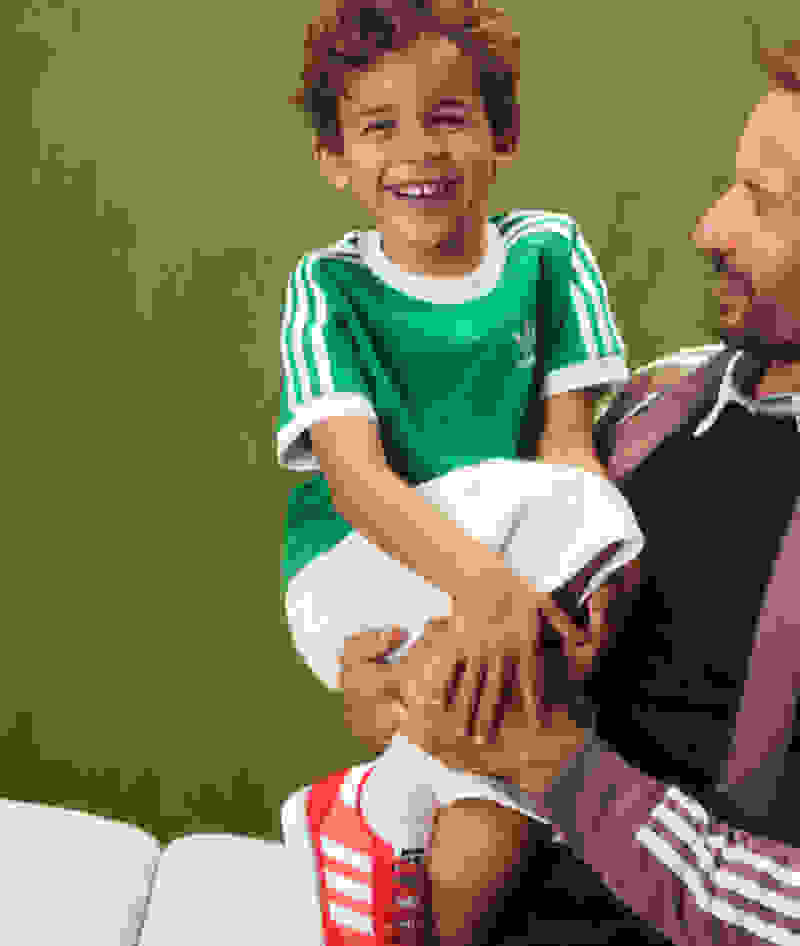A young boy with curly hair wearing a green and white striped shirt is sitting on a couch, smiling brightly, while an adult man with a beard is sitting next to him.