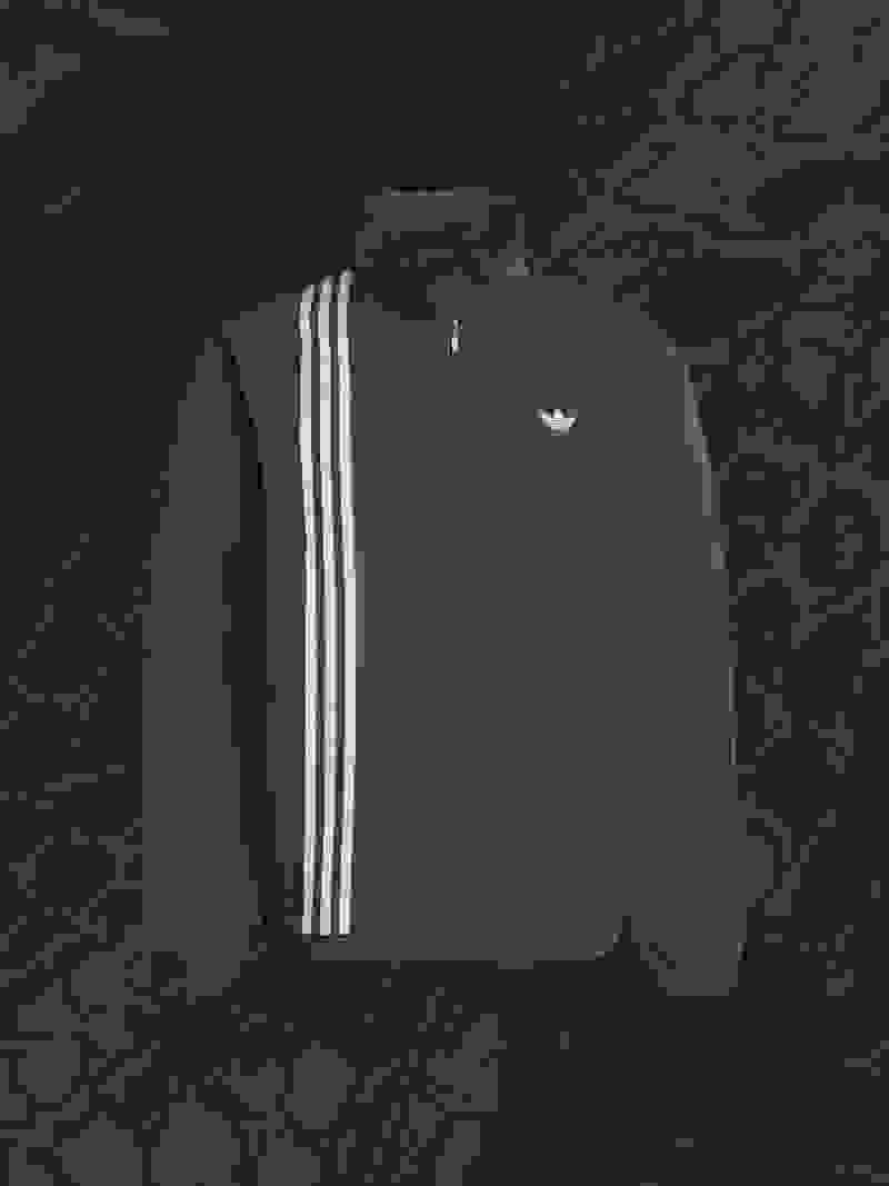 A black Adidas jacket with white stripes stands out against a dark, textured background.
