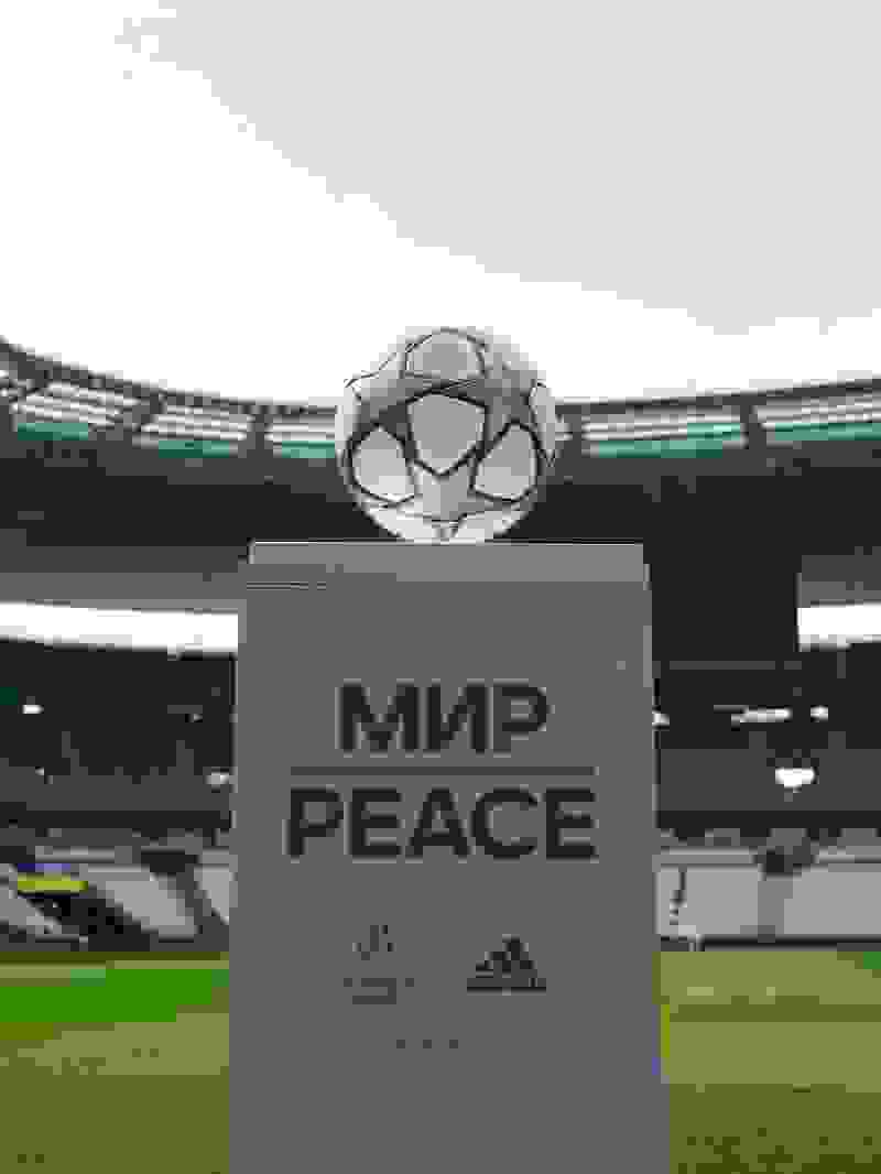 A large soccer ball-shaped object sits atop a pedestal displaying the text "MIR PEACE" in a sports stadium setting with a grassy field and stands visible in the background.