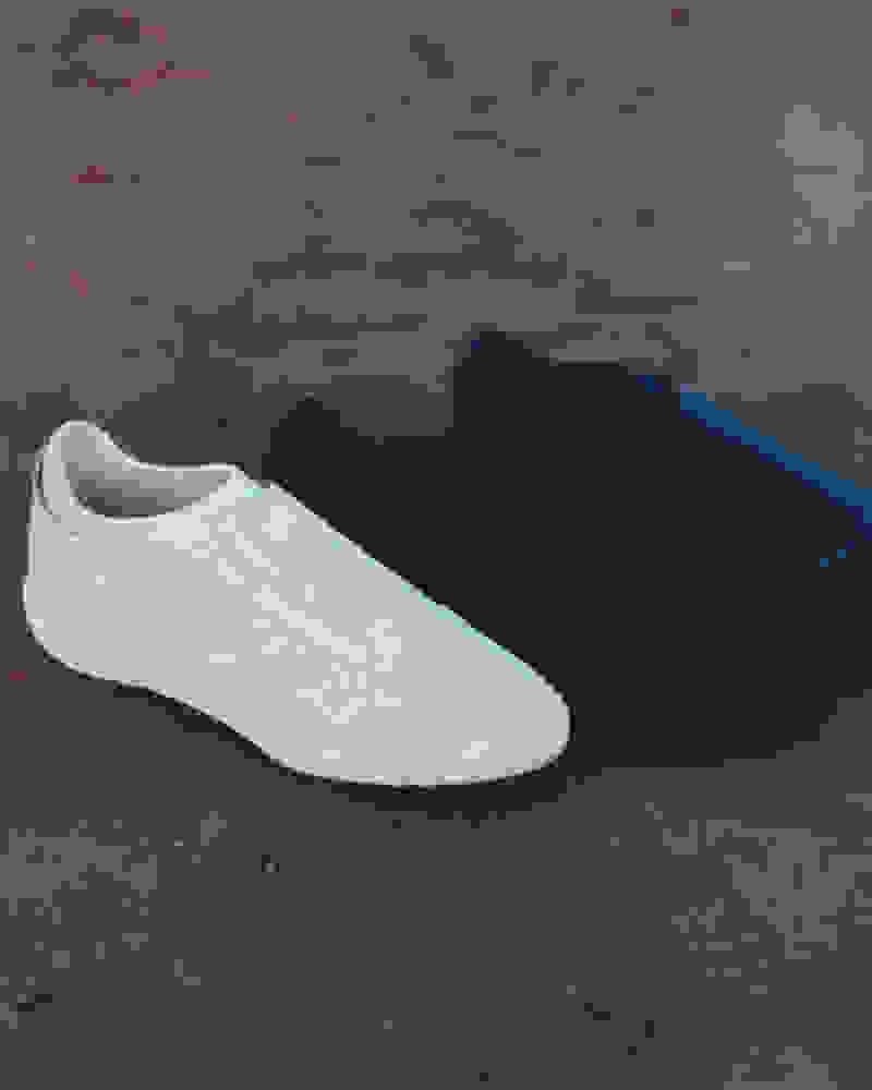 The image shows a pair of casual slip-on shoes, one white and one black, resting on a textured, reddish-brown surface.