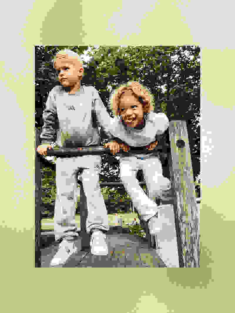 Twee jonge kinderen, een jongen en een meisje, staan op een houten platform in een bebost gebied, omgeven door weelderig groen.