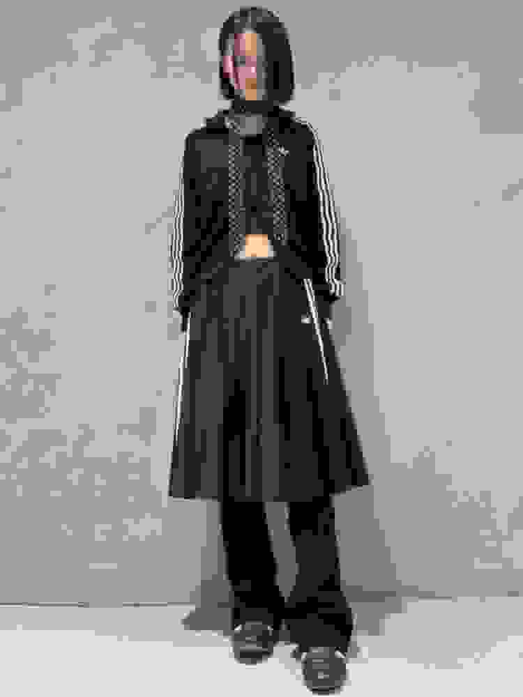 A person wearing a black jacket, crop top, and a long black skirt stands in front of a plain gray background.