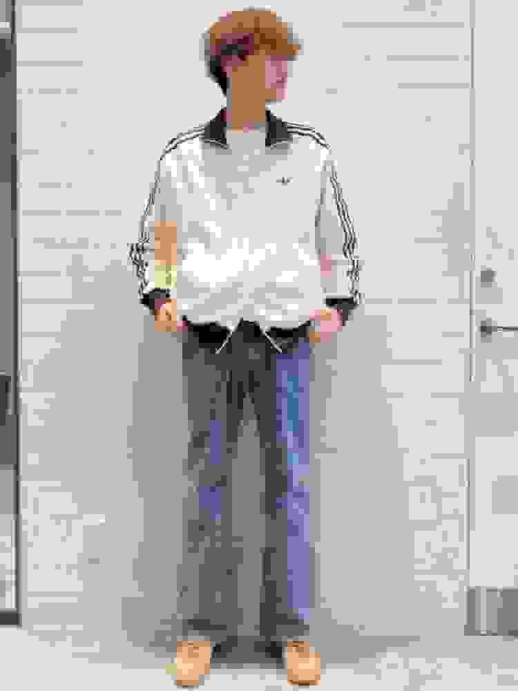 A person wearing a white jacket with black stripes, blue jeans, and beige shoes stands in front of a white wall.