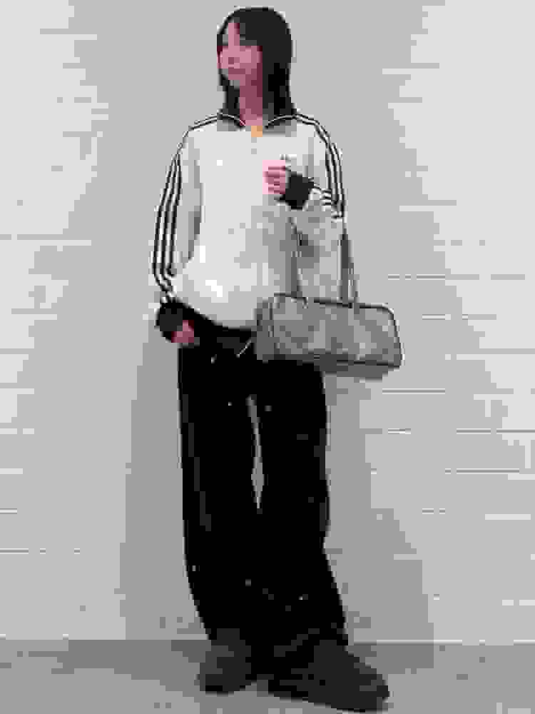 A young woman wearing a beige hooded jacket, black pants, and dark shoes stands in front of a white tiled wall, holding a silver handbag.