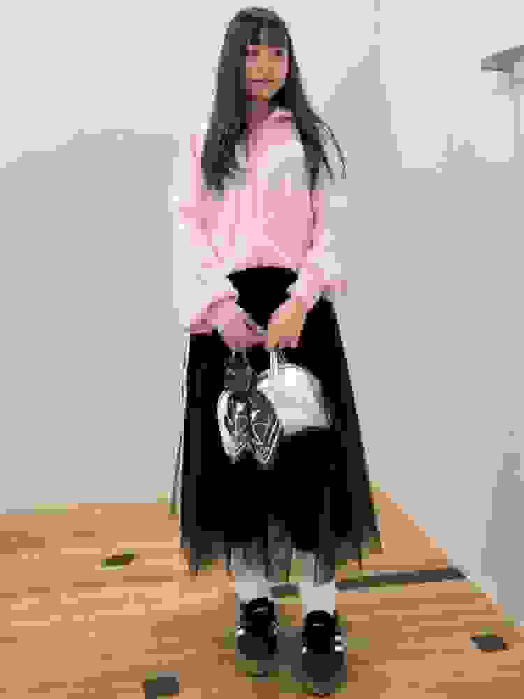 A young person with long dark hair wearing a pink hooded sweatshirt, a black tulle skirt, and black platform shoes stands in a room with a light-colored wall.
