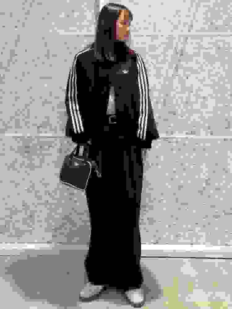 A young woman wearing a black Adidas tracksuit stands in front of a gray concrete wall, holding a black handbag.