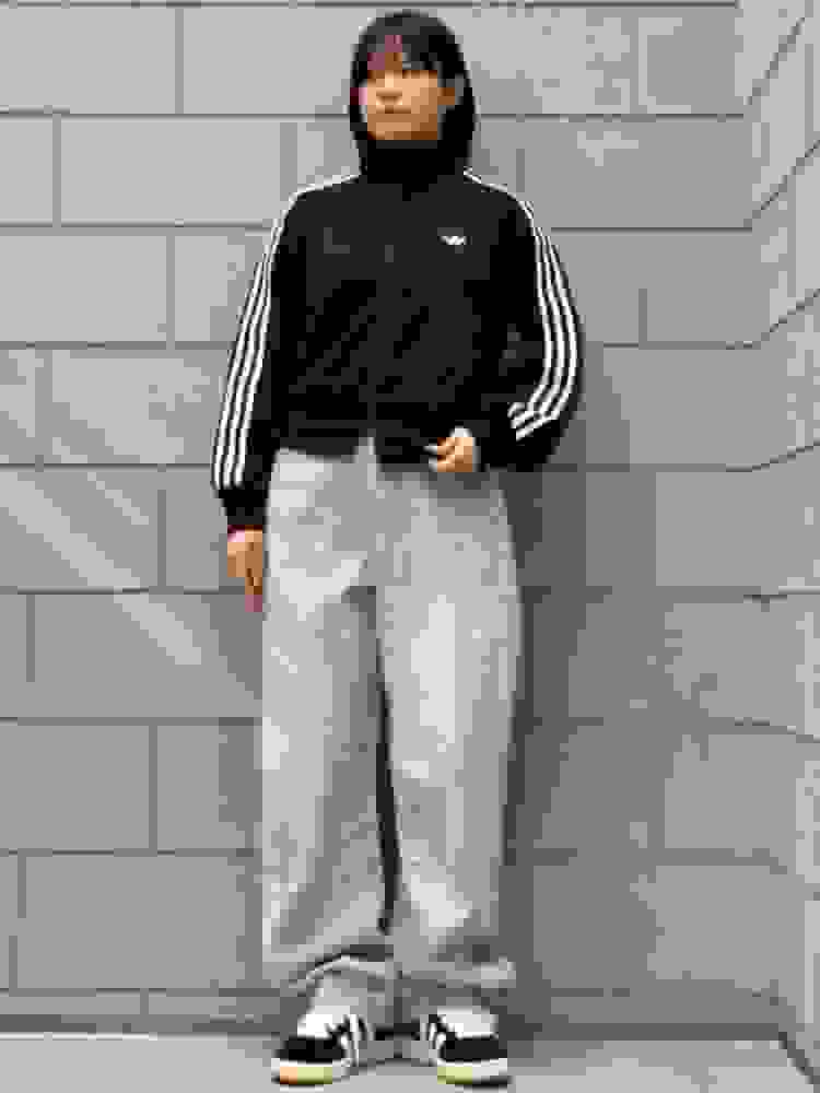 A young person wearing a black Adidas jacket and gray sweatpants stands in front of a gray concrete wall.