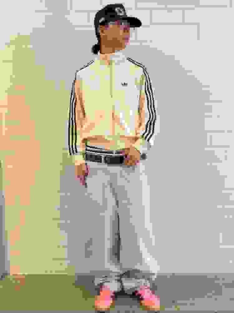 A person wearing a beige jacket with black stripes, a black cap, and pink sneakers stands in front of a plain white wall.