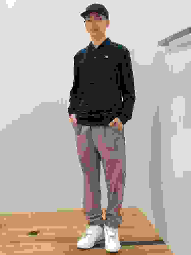 A young man wearing a striped polo shirt, khaki pants, and white sneakers stands in a room with a plain white wall and wooden floor.