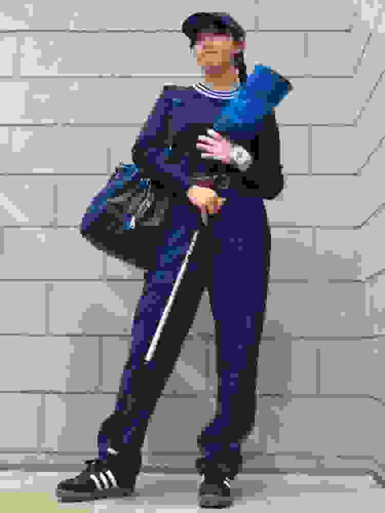 The image shows a person wearing a navy blue outfit, including a sweater and pants, standing in front of a brick wall. They appear to be carrying a bag and have a casual, relaxed posture.