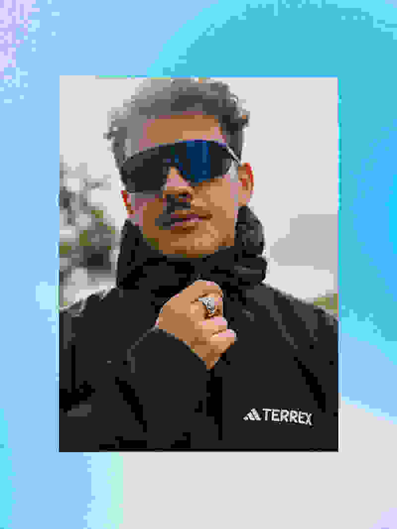 A person wearing a black jacket and sunglasses stands in an outdoor setting with a blue sky in the background.