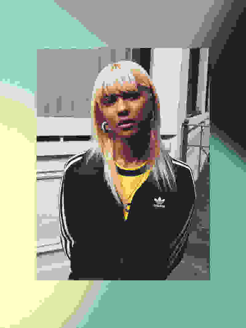 A young woman with blonde hair wearing a black Adidas jacket stands in front of a gray background.