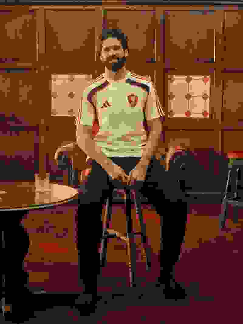 A man with a beard wearing a white and red sports jersey is seated in a wooden chair in a room with ornate wooden paneling and patterned tile flooring.