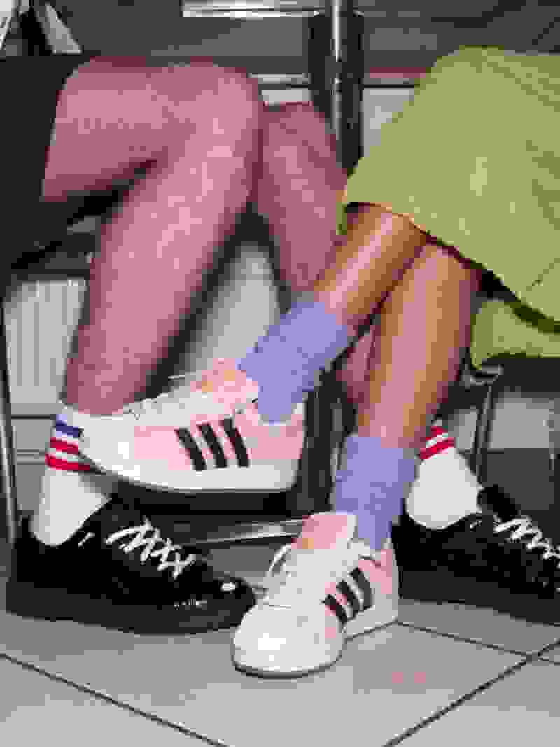 The image shows a person's legs and feet, with the person wearing colorful socks and sneakers, sitting on what appears to be a tiled floor in a room with shelves or cabinets visible in the background.