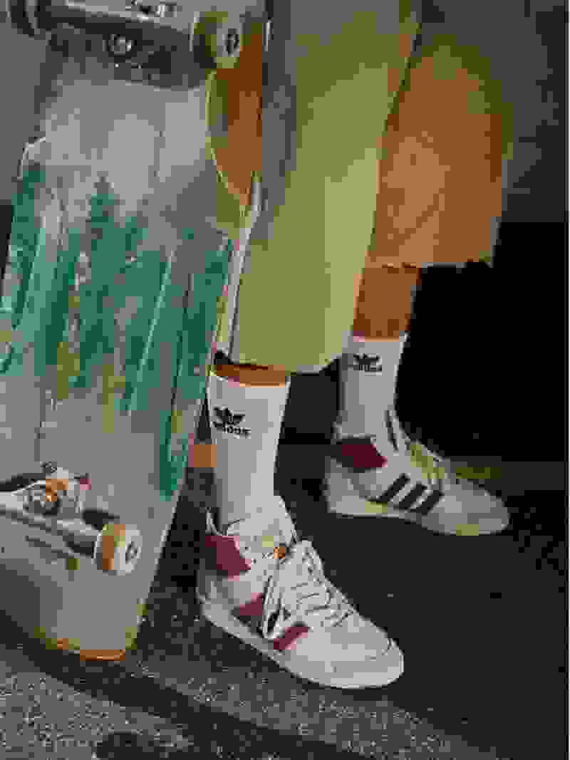 The image shows a person's lower body wearing Adidas sneakers and socks, standing next to a skateboard and a paper bag on a concrete floor.