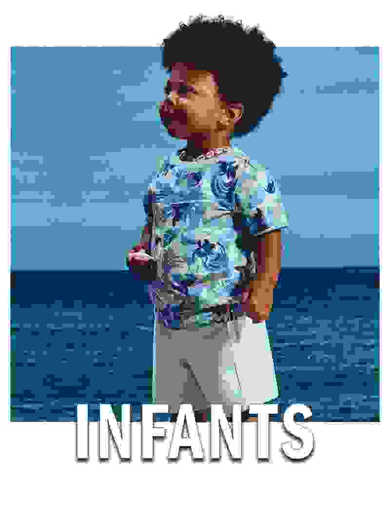 A young child with curly dark hair stands on a beach, wearing a colorful floral-patterned shirt, against the backdrop of a serene blue ocean.